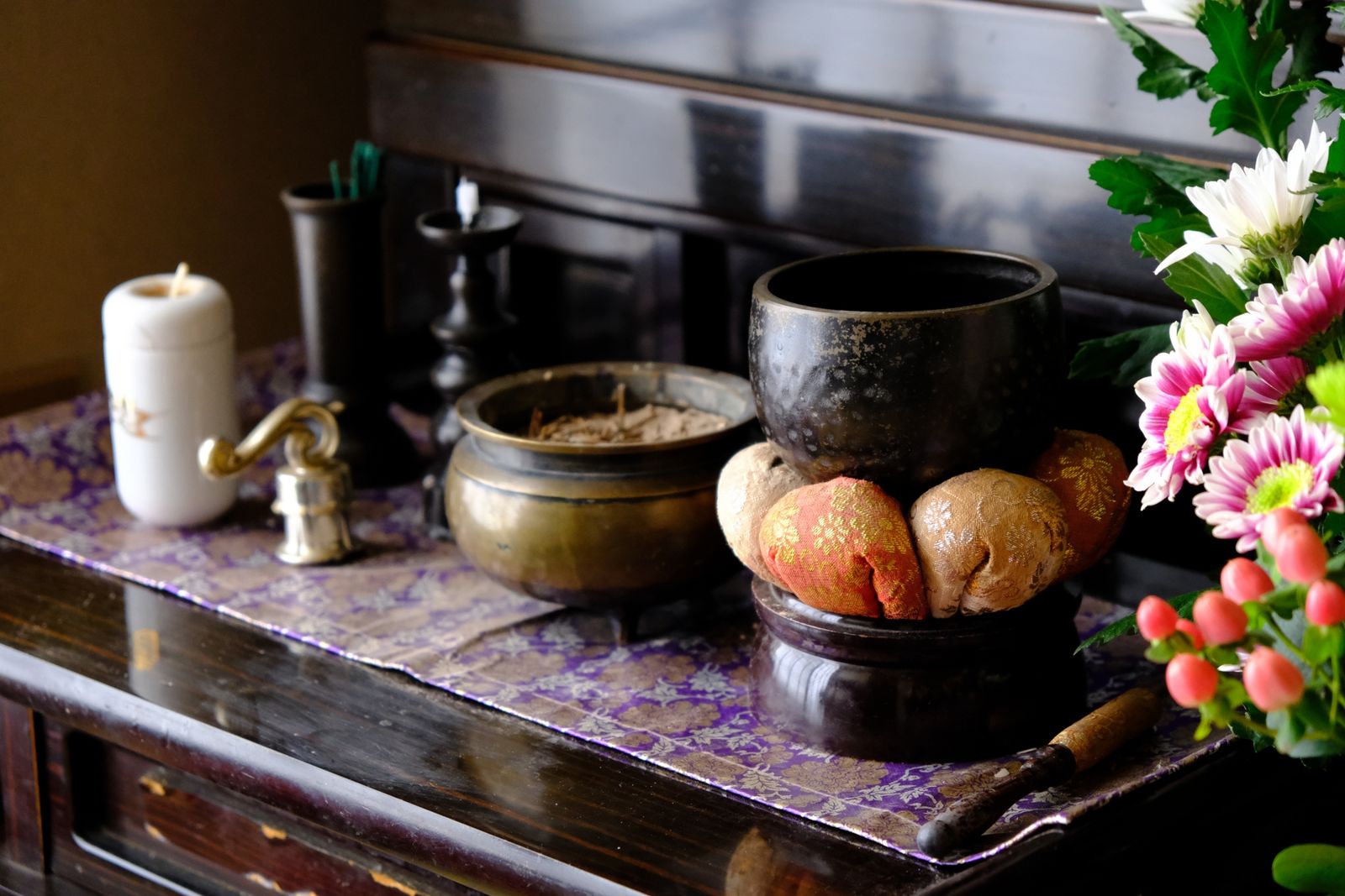 A family butsudan (Buddhist altar) laid out for Obon — an orin bell on its brocade cushion with its striker, a brass incense burner, a white candle, and chrysanthemums with red hypericum berries over a purple-and-gold cloth.