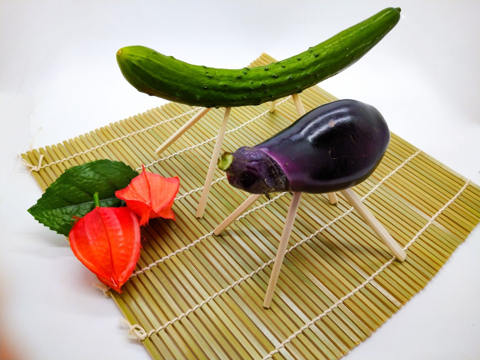 Shoryo-uma: a cucumber horse and an eggplant ox, each standing on legs made of split chopsticks, set on a bamboo mat beside a sprig of red hozuki lantern fruit. The horse is said to bring the spirits home quickly, the ox to carry them back slowly — though the telling varies from place to place.