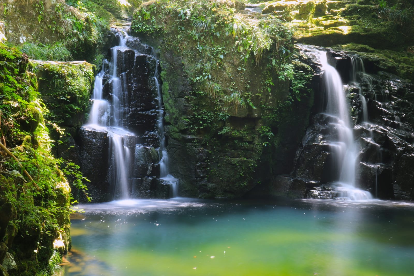 Twin strands sliding down a dark rock face into a clear blue-green pool.