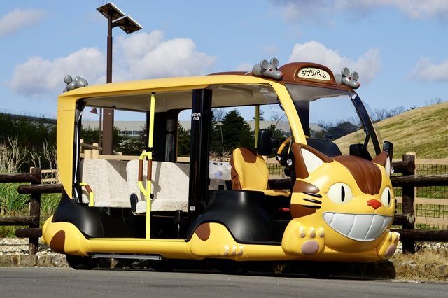 The cheerful yellow Catbus shuttle, grinning beside a wooden fence in the park.