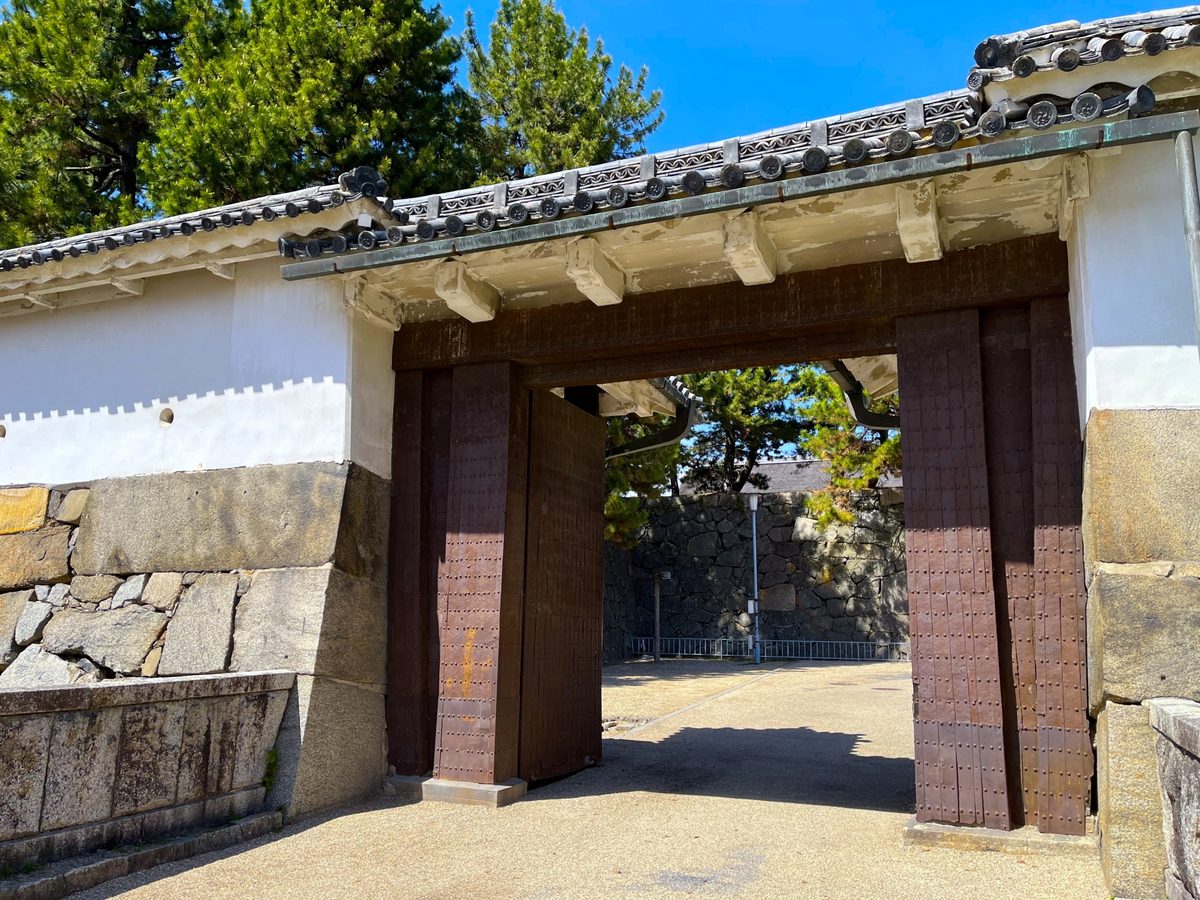 A castle gate with heavy wooden doors set in white plaster walls atop massive stone foundations.