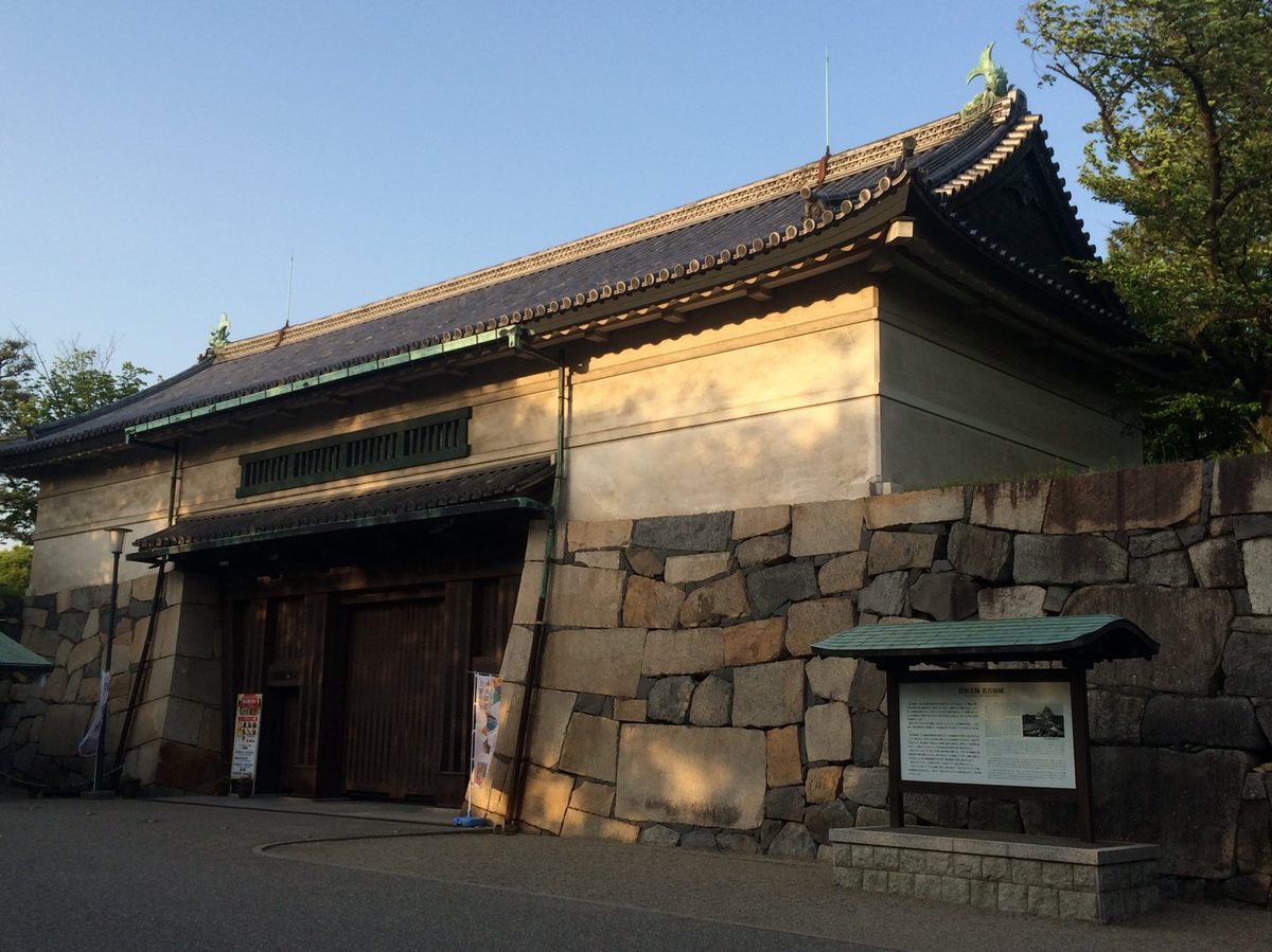 A copper-roofed castle gate at golden hour, a small golden shachihoko on its ridge above the stone wall.