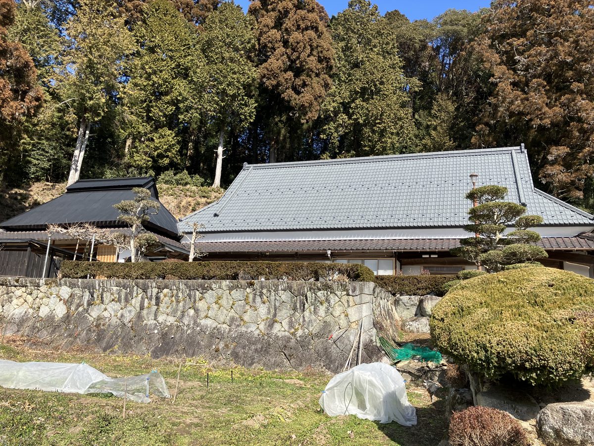 The tiled-roof farmhouse on its stone embankment, backed by wooded hills.