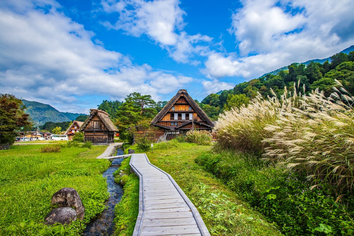 A wooden path along a stream leading to gassho houses in autumn