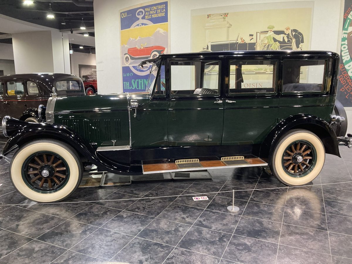 A stately dark-green 1920s sedan on wooden-spoke wheels, set against a wall of vintage motoring posters.