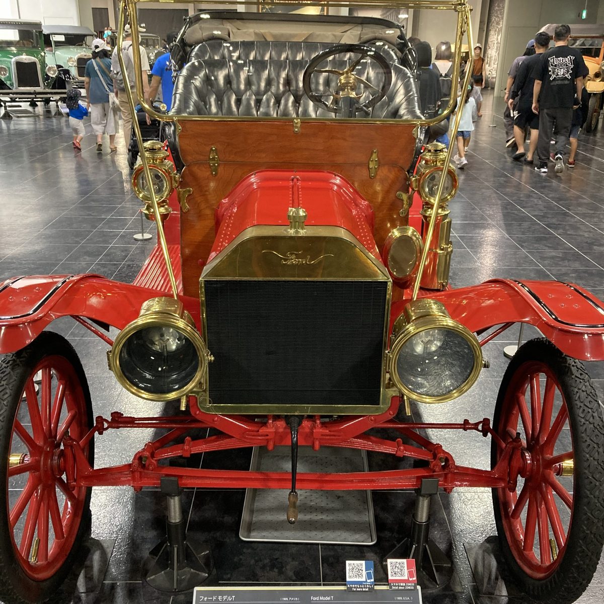 The front of the red 1908 Ford Model T — brass lamps, red spoked wheels, and a crowd gathered round.